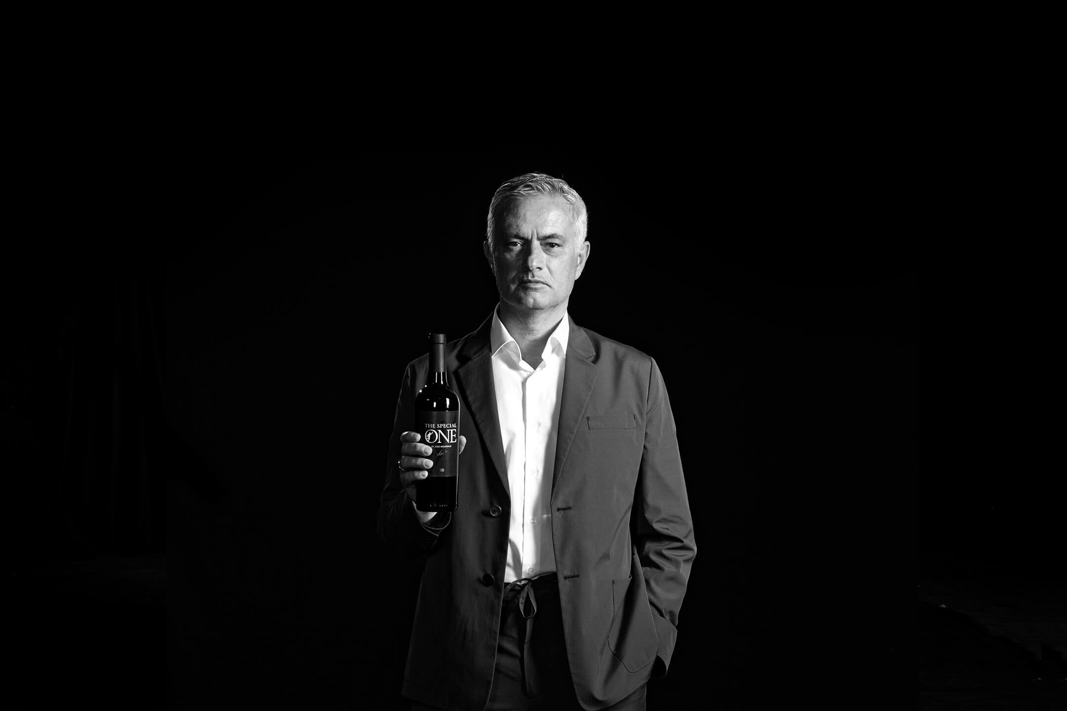 José Mourinho in a suit holding a bottle of "The Special One by José Mourinho" red wine in a dark setting.