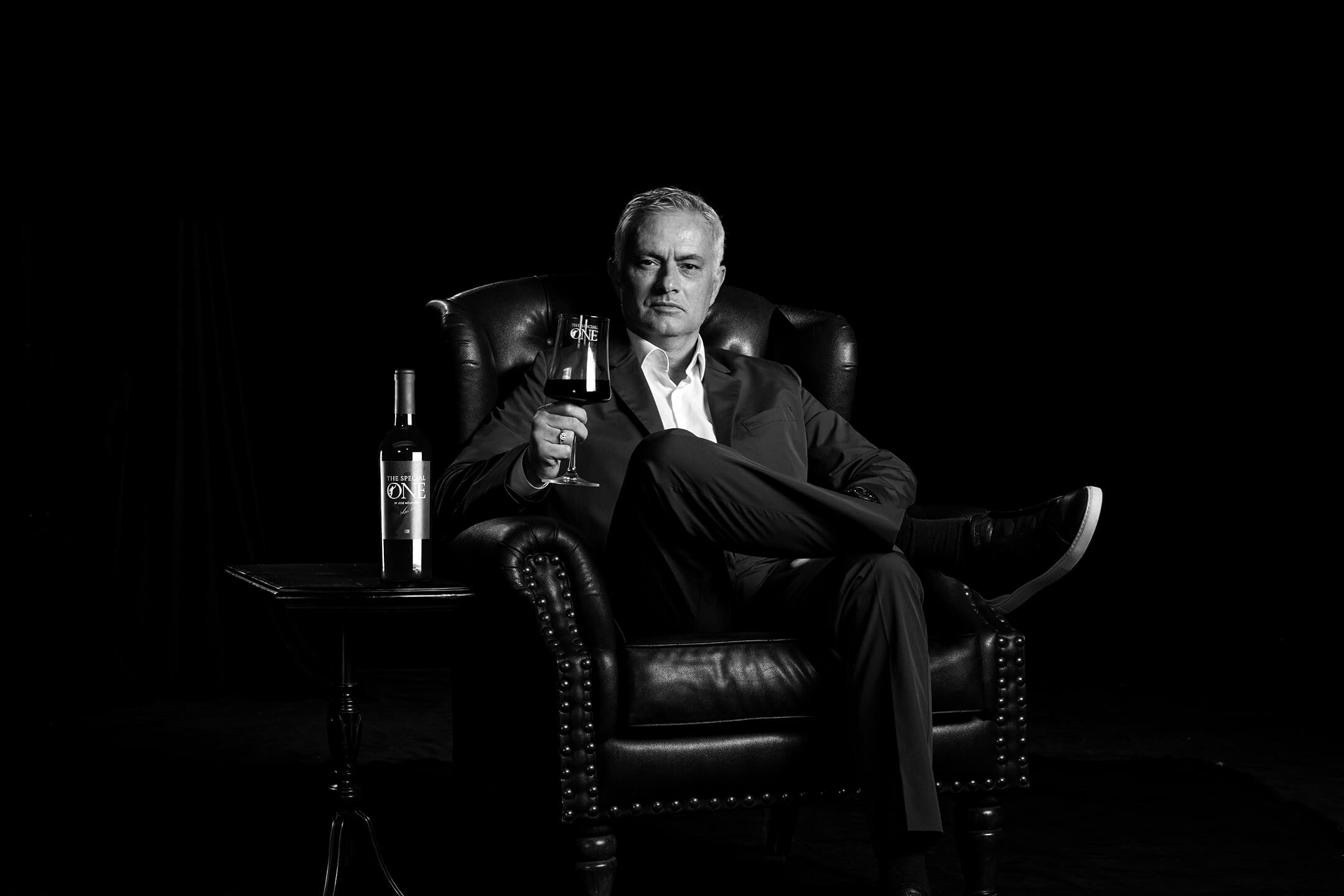 Black and white image of José Mourinho in suit sitting in a leather chair holding a wine glass and a bottle of red wine labeled "The Special One by José Mourinho" on a side table.